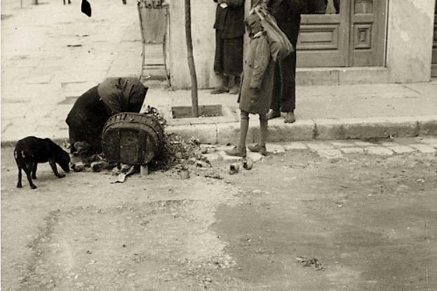 Κατοχή, ο σκληρός χειμώνας 1941-1942. Πειραιάς, στη γωνία Γεωργίου Α΄ και Πραξιτέλους. Μια γυναίκα ψάχνει στα σκουπίδια για φαγητό.